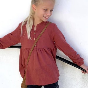 Puede incluir: Una persona joven lleva una camisa de manga larga abotonada de color &oacute;xido con cintura fruncida. La camisa tiene una correa de cuero marr&oacute;n que cuelga del cuello. La persona lleva pantalones negros con bolsillos cargo.