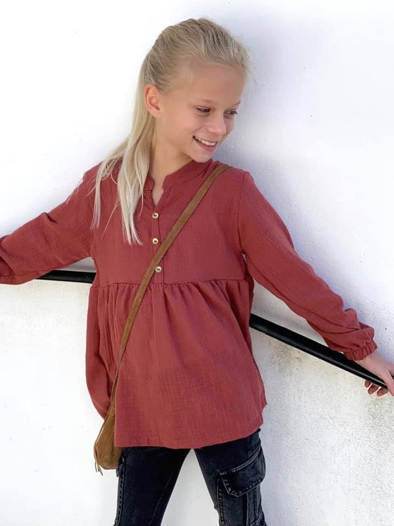 Puede incluir: Una persona joven lleva una camisa de manga larga abotonada de color &oacute;xido con cintura fruncida. La camisa tiene una correa de cuero marr&oacute;n que cuelga del cuello. La persona lleva pantalones negros con bolsillos cargo.