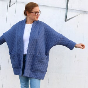 May include: A blue quilted jacket with a diamond pattern, featuring large front pockets and a relaxed fit. The jacket has a shawl collar and long sleeves with turned-up cuffs. The model is wearing a white shirt and blue jeans.