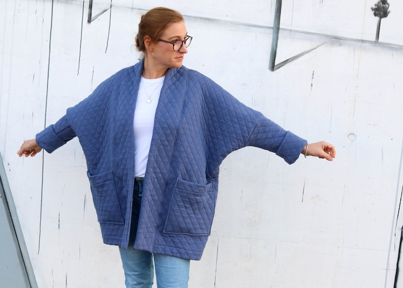 May include: A blue quilted jacket with a diamond pattern, featuring large front pockets and a relaxed fit. The jacket has a shawl collar and long sleeves with turned-up cuffs. The model is wearing a white shirt and blue jeans.