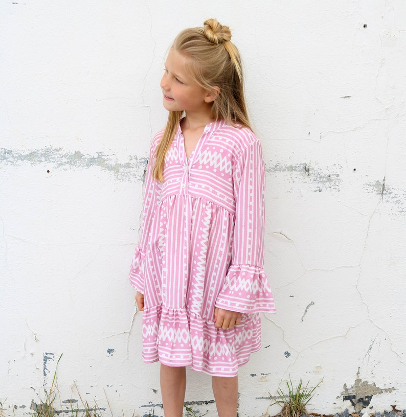 Puede incluir: Un vestido de rayas rosa y blancas con un patr&oacute;n geom&eacute;trico blanco. El vestido tiene mangas largas con pu&ntilde;os acampanados y una falda de capas.