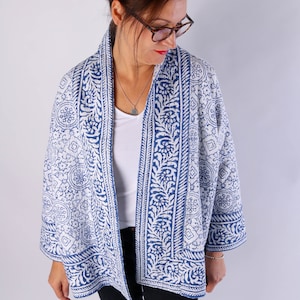 May include: A blue and white patterned kimono jacket. The jacket has a repeating floral and geometric design. It is open-fronted with three-quarter length sleeves. The jacket is worn over a white top and black trousers.