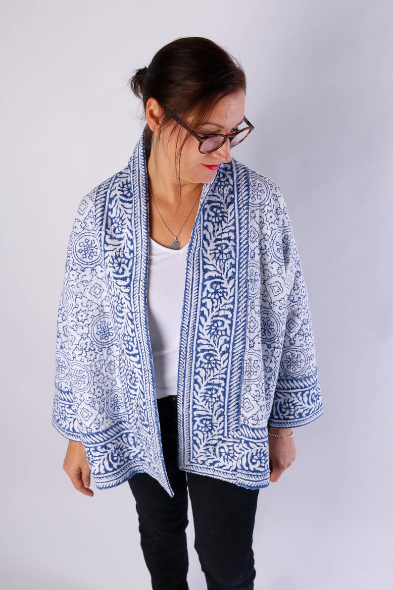 May include: A blue and white patterned kimono jacket. The jacket has a repeating floral and geometric design. It is open-fronted with three-quarter length sleeves. The jacket is worn over a white top and black trousers.