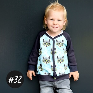 Könnte beinhalten: Ein Kind trägt einen blau-schwarzen Cardigan mit einem Waschbär-Druck. Der Cardigan hat einen Knopfverschluss und einen grünen Besatz. Das Kind trägt eine graue Jeans.
