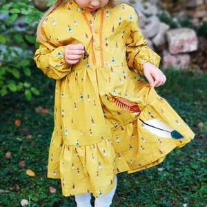 Puede incluir: Un vestido amarillo de manga larga y dobladillo con volantes. El vestido est&aacute; cubierto de un patr&oacute;n de plumas de colores.