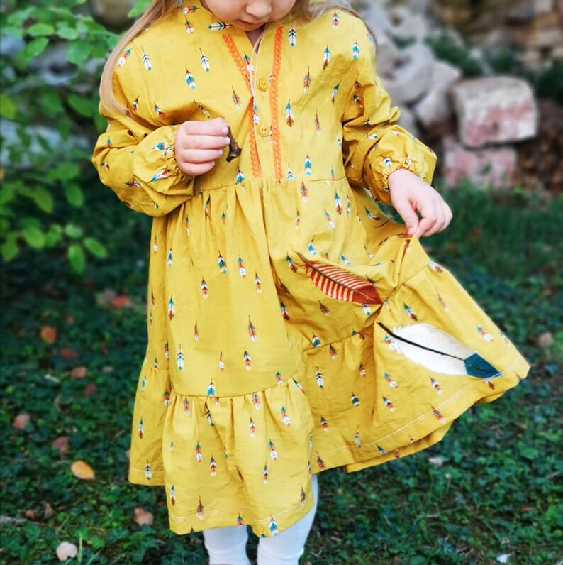 Puede incluir: Un vestido amarillo de manga larga y dobladillo con volantes. El vestido est&aacute; cubierto de un patr&oacute;n de plumas de colores.