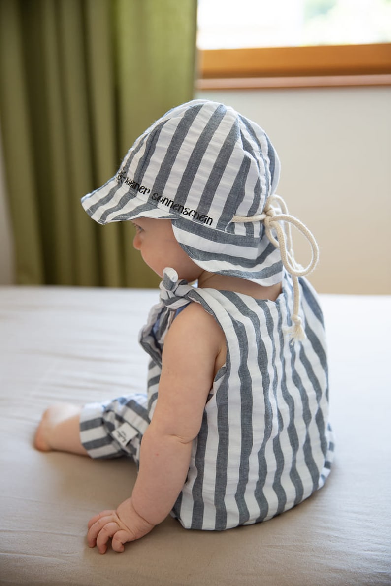May include: A baby wearing a blue and white striped sun hat and romper. The hat has a brim and ties, with the text "kleiner sonnenschein" printed on the brim. The romper has a sleeveless design.