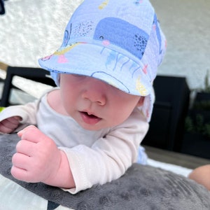May include: A light blue baby sun hat with a whale and sea creature pattern. The hat has a brim and is worn by a baby. The baby is wearing a white long-sleeved shirt and is lying on a gray pillow.