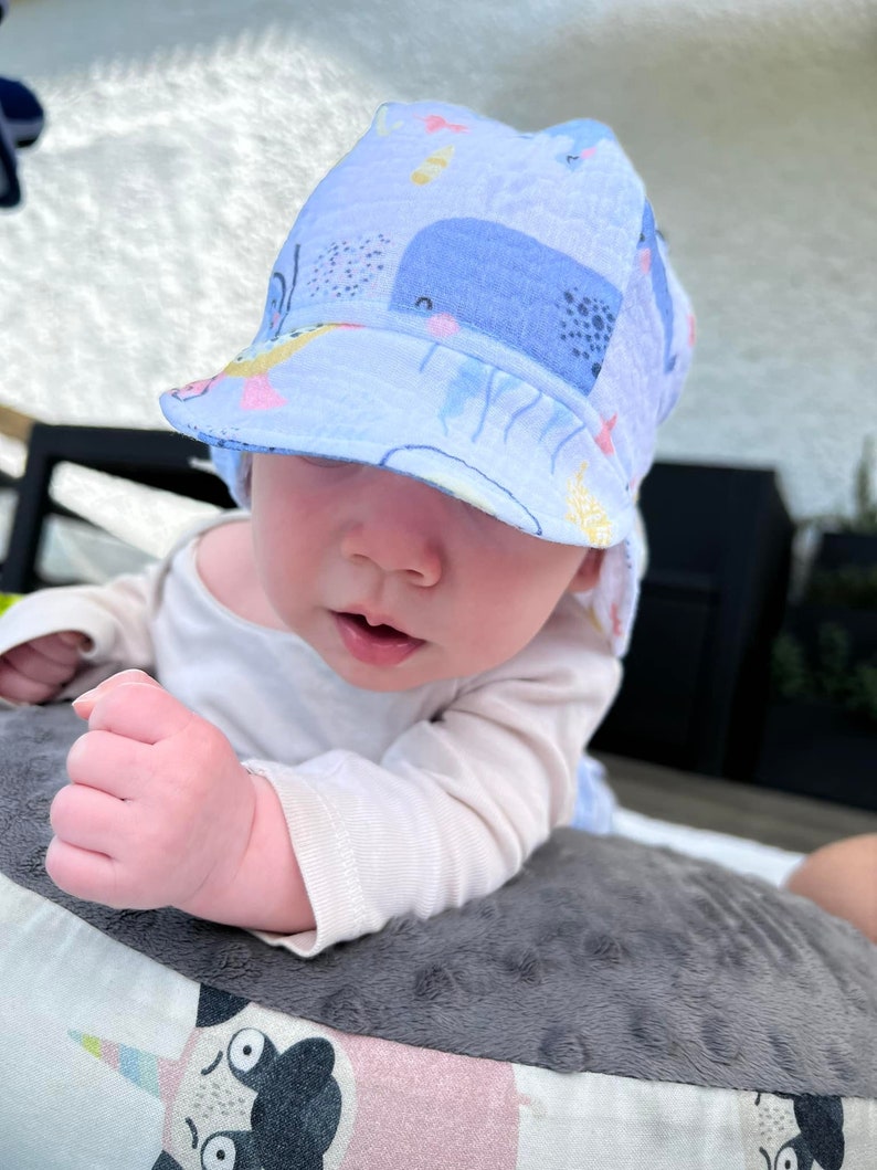 May include: A light blue baby sun hat with a whale and sea creature pattern. The hat has a brim and is worn by a baby. The baby is wearing a white long-sleeved shirt and is lying on a gray pillow.