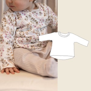 Puede incluir: Una camisa blanca de manga larga con cuello redondo. La camisa tiene un dobladillo trasero ligeramente más largo que el delantero. La camisa se muestra en un bebé que lleva una camisa y pantalones con estampado floral.