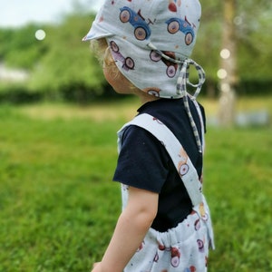 May include: A light blue baby sun hat, overalls, and shorts set. The set features a repeating pattern of blue and red toy cars. The hat has a brim and ties. The child is wearing a black t-shirt underneath the overalls.