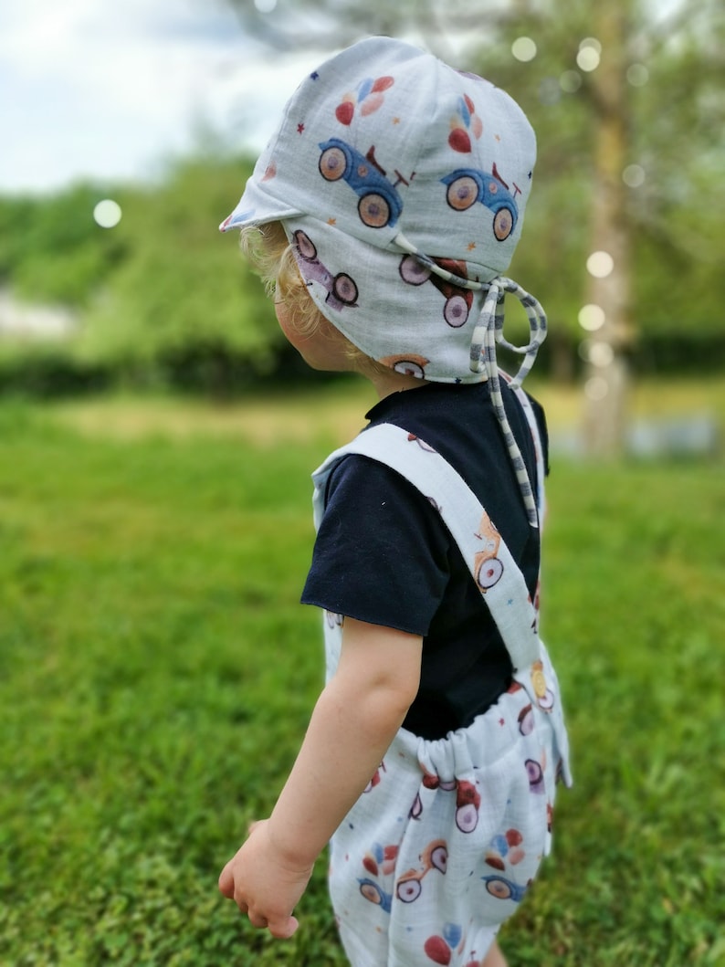 May include: A light blue baby sun hat, overalls, and shorts set. The set features a repeating pattern of blue and red toy cars. The hat has a brim and ties. The child is wearing a black t-shirt underneath the overalls.