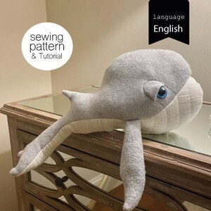 May include: A gray whale plush toy with a white belly and blue eyes. The whale is lying on a wooden table with a mirrored surface. The image includes the text "sewing pattern & Tutorial" and "language English".