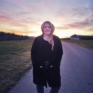 May include: A person wearing a black coat, black top, and blue jeans stands on a paved path. The background shows a sunset with orange and pink colours, a field, and a distant building.