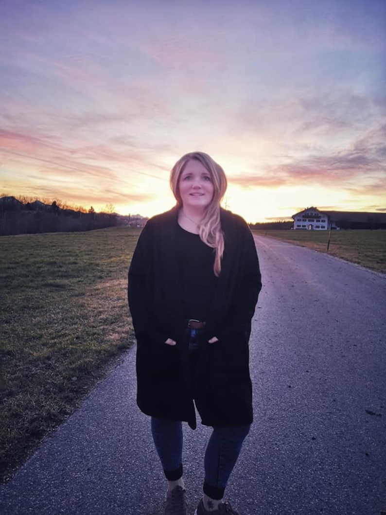 May include: A person wearing a black coat, black top, and blue jeans stands on a paved path. The background shows a sunset with orange and pink colours, a field, and a distant building.