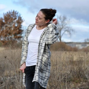 May include: A woman in a white t-shirt, black jeans, and a long, open plaid jacket in cream and black. The jacket has large pockets. The background shows a field and a cloudy sky.