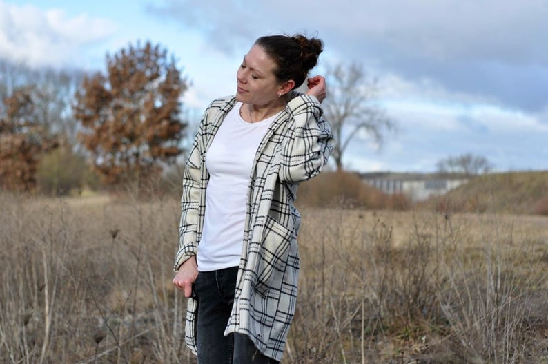 May include: A woman in a white t-shirt, black jeans, and a long, open plaid jacket in cream and black. The jacket has large pockets. The background shows a field and a cloudy sky.