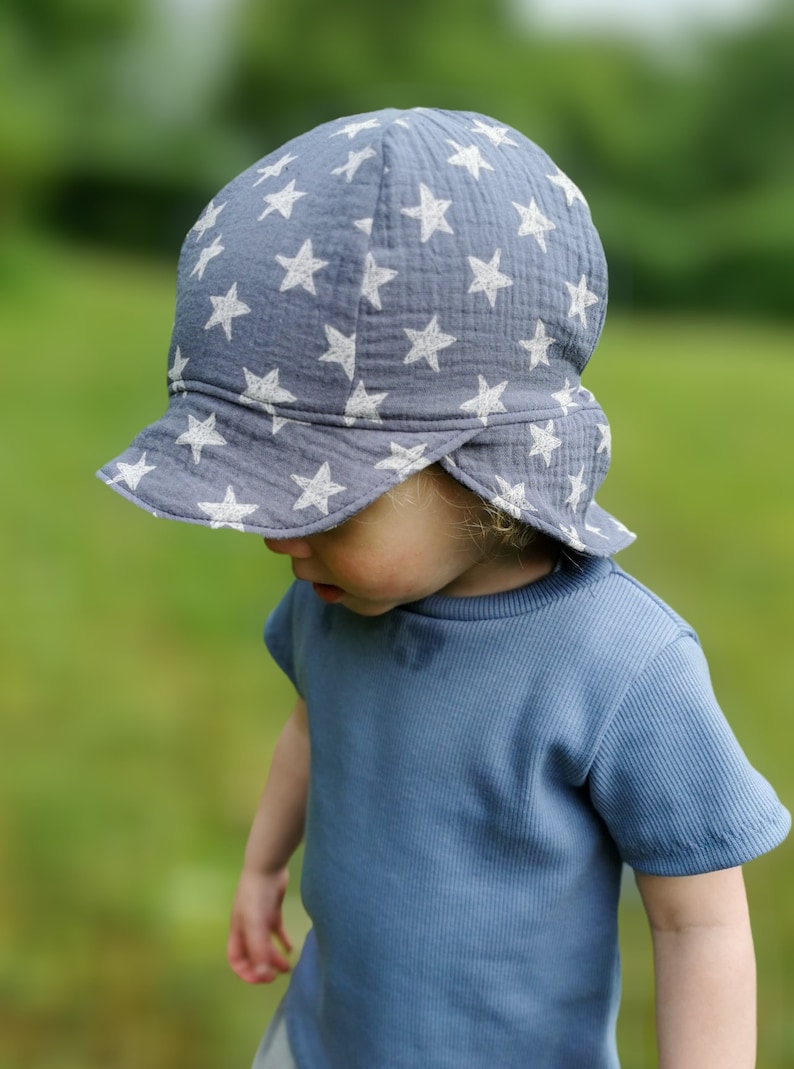May include: A blue sun hat with white star patterns. The hat has a brim and is made of a soft, textured fabric. The child is also wearing a blue short-sleeved shirt.