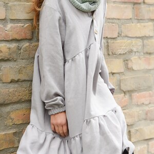 Puede incluir: Un vestido gris de manga larga con un dobladillo con volantes y un frente abotonado. El vestido tiene un ajuste holgado y est&aacute; hecho de una tela suave y c&oacute;moda. La modelo lleva una bufanda de punto verde.