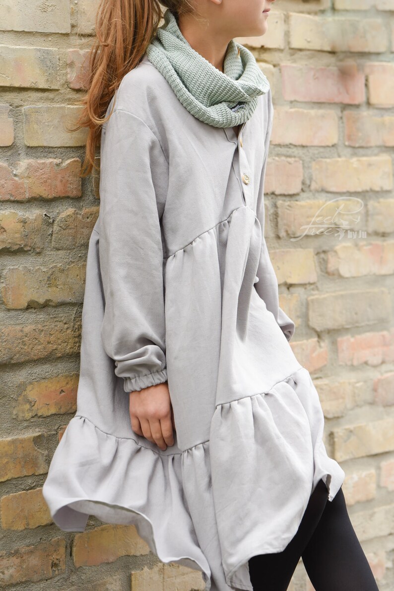 Puede incluir: Un vestido gris de manga larga con un dobladillo con volantes y un frente abotonado. El vestido tiene un ajuste holgado y est&aacute; hecho de una tela suave y c&oacute;moda. La modelo lleva una bufanda de punto verde.