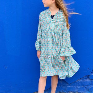 Puede incluir: Una persona joven lleva un vestido azul claro con estampado floral. El vestido tiene mangas largas y un dobladillo con volantes. La persona est&aacute; de pie frente a una pared azul y tiene el pelo recogido en un mo&ntilde;o.