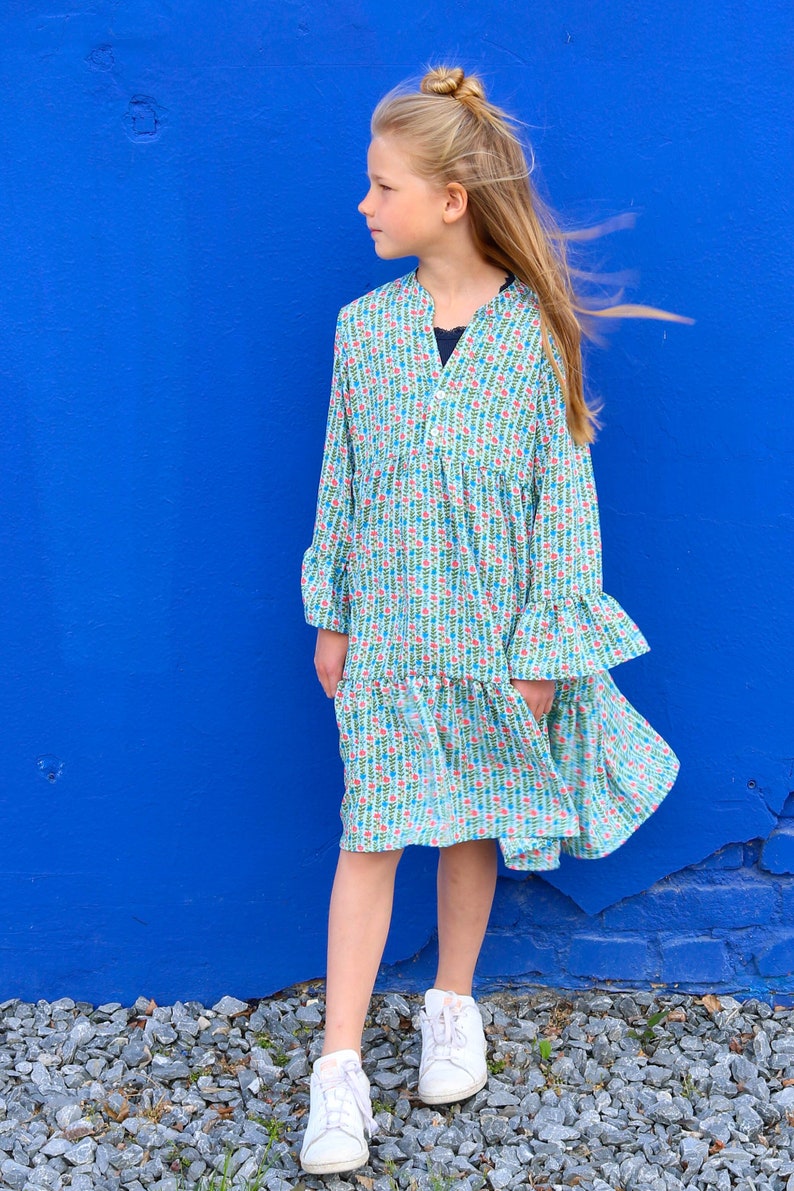 Puede incluir: Una persona joven lleva un vestido azul claro con estampado floral. El vestido tiene mangas largas y un dobladillo con volantes. La persona est&aacute; de pie frente a una pared azul y tiene el pelo recogido en un mo&ntilde;o.