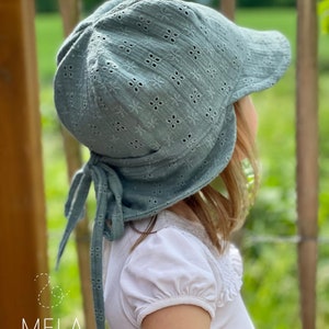 May include: A sage green baby bonnet with a floral eyelet pattern and a brim. The bonnet has ties and is worn by a child. The text "& MELA by Melanie Gieske" is in the lower left corner.