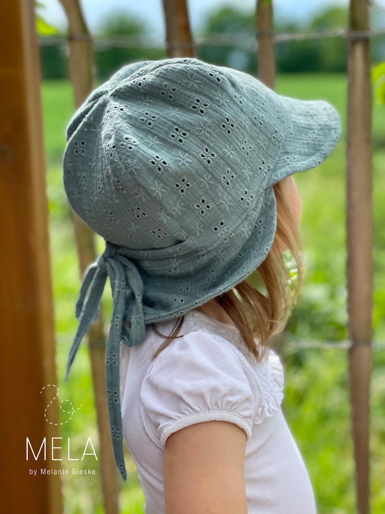 May include: A sage green baby bonnet with a floral eyelet pattern and a brim. The bonnet has ties and is worn by a child. The text "& MELA by Melanie Gieske" is in the lower left corner.