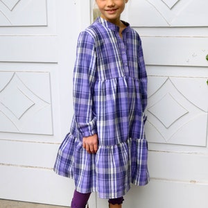 Puede incluir: Una ni&ntilde;a peque&ntilde;a con un vestido de cuadros morados y blancos con un dobladillo con volantes. Lleva botas negras y tiene el pelo recogido en un mo&ntilde;o.