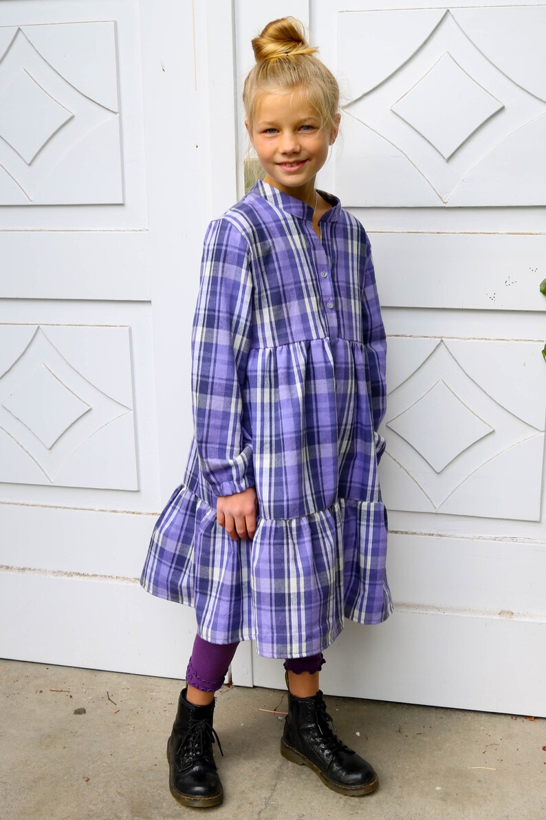 Puede incluir: Una ni&ntilde;a peque&ntilde;a con un vestido de cuadros morados y blancos con un dobladillo con volantes. Lleva botas negras y tiene el pelo recogido en un mo&ntilde;o.