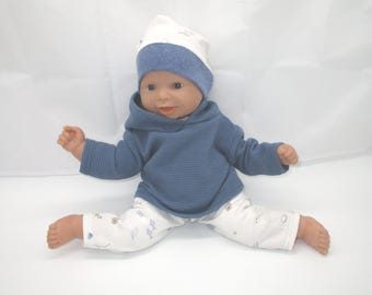 May include: A baby doll wearing a blue hooded sweatshirt, a white and blue beanie, and white pants with a blue pattern. The doll is posed with its legs spread apart.