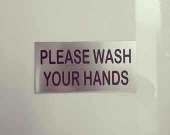 May include: A silver sign with black text that reads "Please wash your hands".