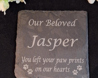 Może przedstawiać: Kwadratowa, szara tablica pamiątkowa z łupku z napisem "Our Beloved Jasper". Poniżej napis "You left your paw prints on our hearts" z grafiką odcisków łap. Białe kwiaty w tle.