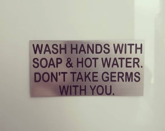 May include: A silver sign with black text that reads "Wash hands with soap & hot water. Don't take germs with you."