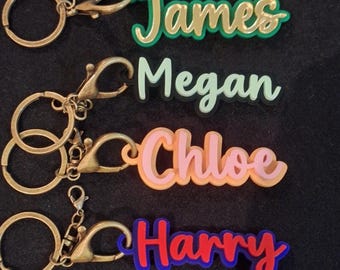 May include: Four personalized keychains with names: James, Megan, Chloe, and Harry. Each name is in a different color scheme, with a gold-colored keychain ring and clasp. The names are in a layered, 3D style, with a variety of colors.