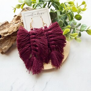 May include: A pair of deep burgundy tassel earrings featuring a knotted macrame design in a leaf shape. The earrings are attached to a white display card with the text "nunture designs" and have gold-toned metal hooks for attachment.
