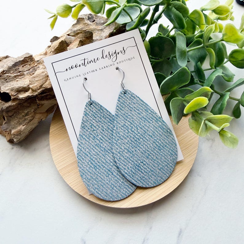May include: A pair of light blue teardrop earrings with a textured pattern, displayed on a wooden stand. The earrings are hanging from silver hooks. A white card with the text "noontime designs" is behind the earrings.