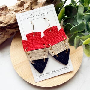 May include: A pair of layered dangle earrings featuring three stacked geometric shapes in red, beige, and black textured material. The pieces are connected by gold-tone metal jump rings and hang from gold-tone hooks on a white display card with the text Noontime Designs Jewelry Boutique and WWW.NOONTIMEDESIGNS.COM.