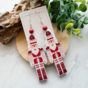 May include: A pair of dangle earrings featuring a festive nutcracker design. The earrings are primarily white and red, with glitter accents. Each earring has a red bead at the top and a silver hook. The earrings are displayed on a white card with the text "nuntime designs".