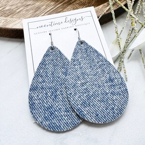 May include: Teardrop earrings with a blue and white denim effect. The earrings are displayed on a white card with the text "noontime designs GENUINE LEATHER EARRING BOUTIQUE". The earrings have silver-coloured hooks. The earrings are a stylish accessory.