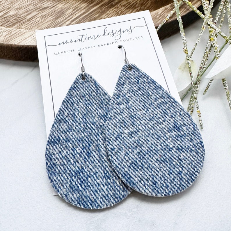 May include: Teardrop earrings with a blue and white denim effect. The earrings are displayed on a white card with the text "noontime designs GENUINE LEATHER EARRING BOUTIQUE". The earrings have silver-coloured hooks. The earrings are a stylish accessory.