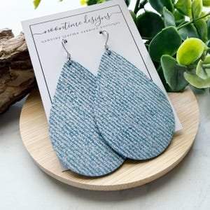 May include: A pair of blue and white teardrop-shaped earrings with a textured pattern. The earrings are displayed on a white card with the text "noontime designs GENUINE LEATHER LARRING BOUTIQUE". The earrings are on a wooden tray.