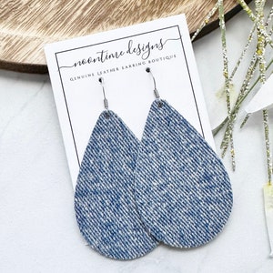 May include: Teardrop earrings with a denim-style texture, suspended from silver hooks. The earrings are presented on a white card with the text "noontime designs GENUINE LEATHER EARRING BOUTIQUE". The background features a wooden tray and decorative sprigs.