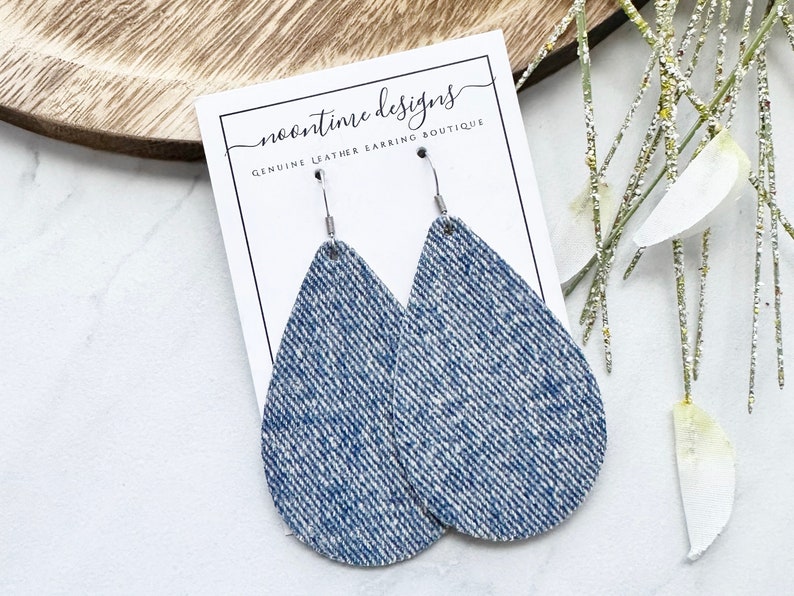May include: Teardrop earrings with a denim-style texture, suspended from silver hooks. The earrings are presented on a white card with the text "noontime designs GENUINE LEATHER EARRING BOUTIQUE". The background features a wooden tray and decorative sprigs.