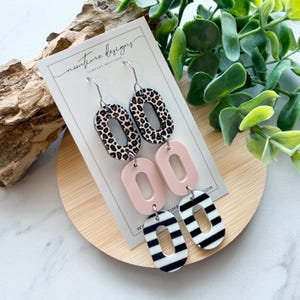 May include: Dangle earrings with a layered design. The top oval features a leopard print, followed by a light pink oval, and a black and white striped oval at the bottom. The earrings are displayed on a white card with the text "noontime designs JEWELRY BOUTIQUE".