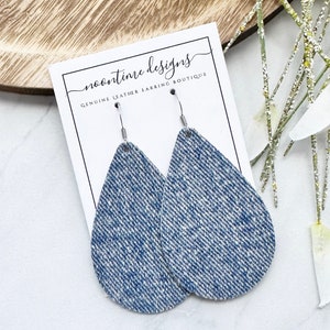 May include: Teardrop earrings with a denim effect, featuring silver hooks. The earrings are presented on a white card with the text "noontime designs GENUINE LEATHER EARRING BOUTIQUE". The background includes a wooden tray and decorative sprigs.