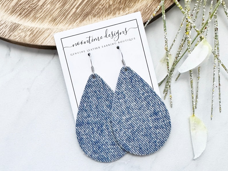 May include: Teardrop earrings with a denim effect, featuring silver hooks. The earrings are presented on a white card with the text "noontime designs GENUINE LEATHER EARRING BOUTIQUE". The background includes a wooden tray and decorative sprigs.