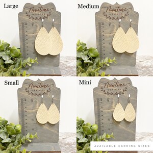 May include: Four pairs of cream-coloured teardrop earrings in various sizes: Large, Medium, Small, and Mini. Each pair hangs from a silver hook and is displayed on a grey wooden stand with a ruler. The stands are labelled with the earring size.