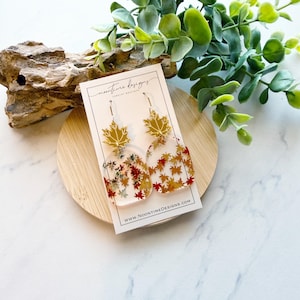 May include: Clear acrylic earrings with gold maple leaf tops and a clear, rounded rectangular body filled with autumn-colored leaves. The earrings are displayed on a white card with the text "noontime designs" and a website address.