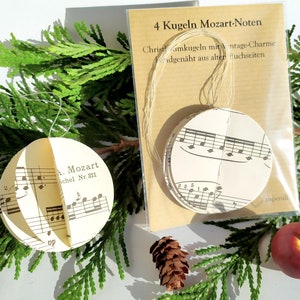 Può includere: Addobbi natalizi realizzati con spartiti musicali vintage. Un ornamento è aperto, rivelando note musicali e il testo "A. Mozart, Schel Nr. 311". La confezione recita "4 Kugeln Mozart-Noten" e "Christbaumkugeln mit vintage-Charme".